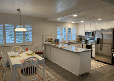 Modern kitchen and dining area with white cabinets, stainless steel appliances, and a set dining table for two. Marble countertops and large windows with shutters are visible.