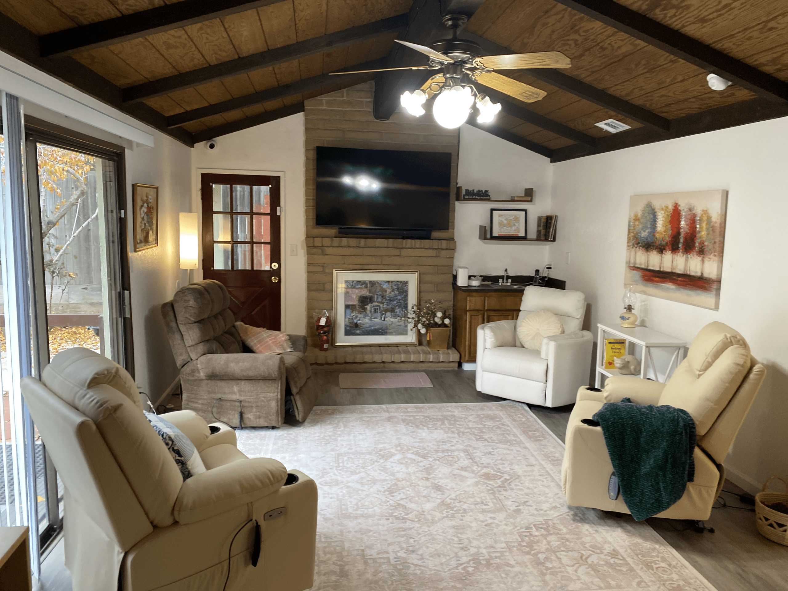 A cozy living room with three recliners, a TV mounted above a fireplace, a ceiling fan, wall art, and sliding glass doors leading outside.