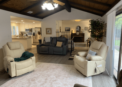 A spacious living room with beige recliners, a dark loveseat, a piano in the dining area, and a person walking in the kitchen. Ceiling fan and large windows provide natural light.