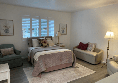 A neatly made bed with decorative pillows sits in a cozy bedroom with a green armchair, a beige loveseat, lamps, artwork, and a window with white shutters.