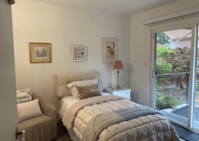 A small, neatly made bedroom with a single bed, armchair, framed art on the wall, a nightstand with a lamp, and a sliding glass door leading to a garden.