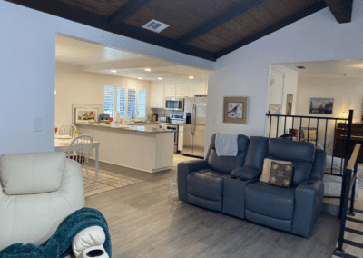 Open living area with gray seating, kitchen with white cabinets and stainless steel appliances, dining table, and wood-look flooring under a dark-beamed ceiling.